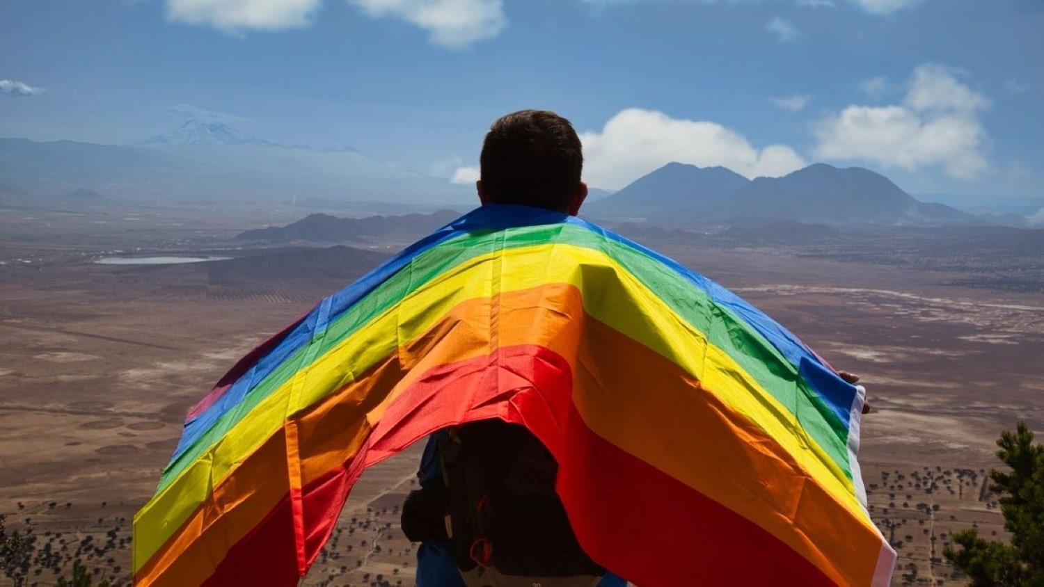 A person holding Pride Flag looking out at a mountain range.